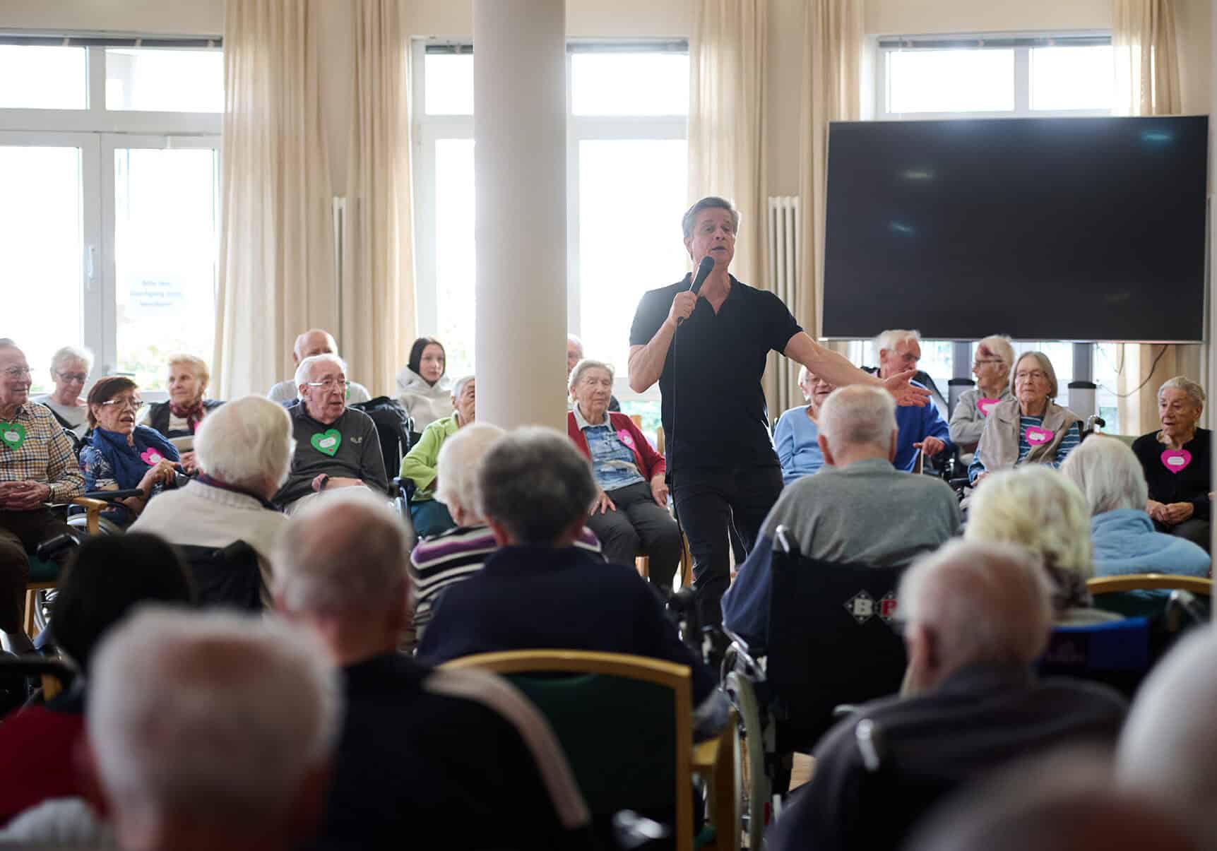 Ein Mann singt vor einer großen Gruppe älterer Menschen in einem Gemeinschaftsraum unserer Residenz "Rethen an der Leine".