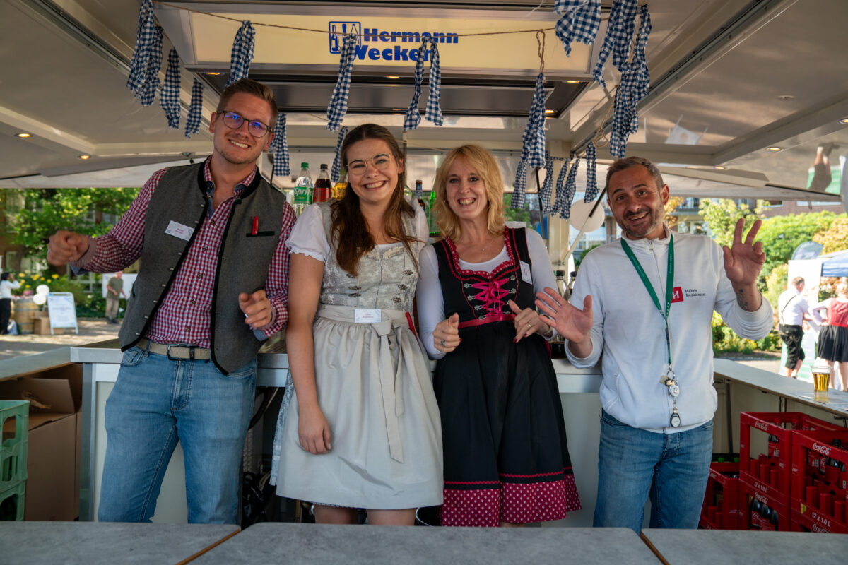 Es werden 4 fröhliche Mitarbeiter vom Schankwagen beim "WiesnZauber" abgebildet. Links steht ein Mann, in der Mitte stehen zwei Frauen und rechts steht ein Mann.